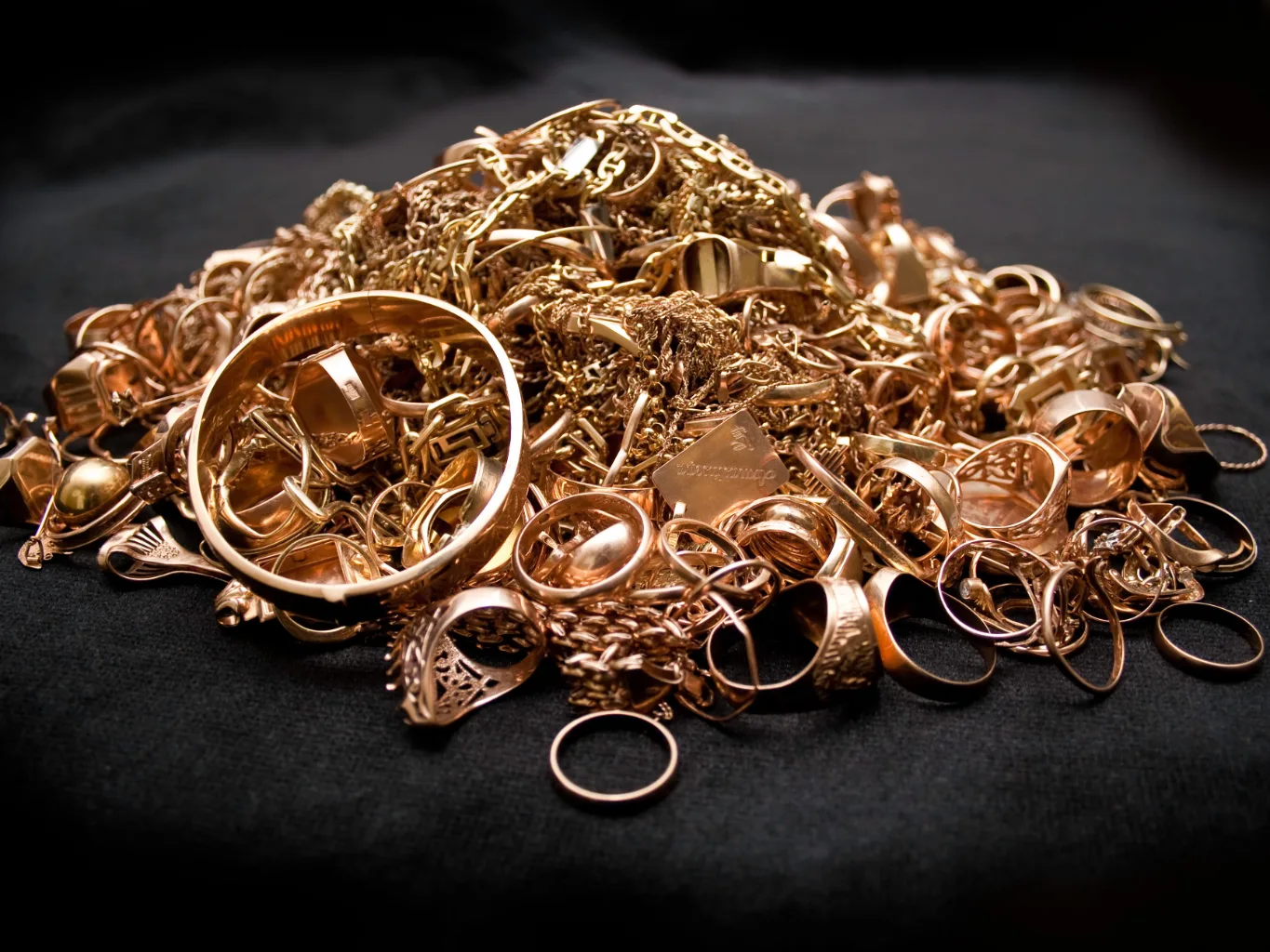 During the intake assessment, a worker uses jeweler's calipers to examine the width of a broken gold chain link on a small tray, with the damaged clasp and loose links beside it