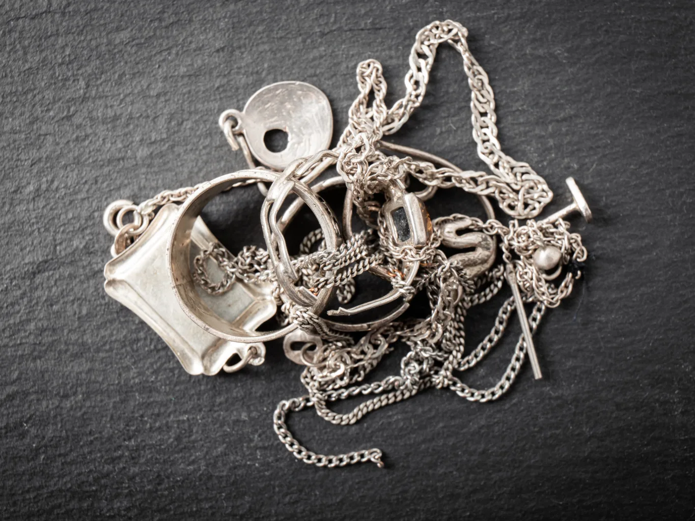 During the intake stage for a broken jewelry purchase, a worker uses chain-nose jewelry pliers to open a damaged clasp over a divided tray containing broken chains, single earrings and bent rings