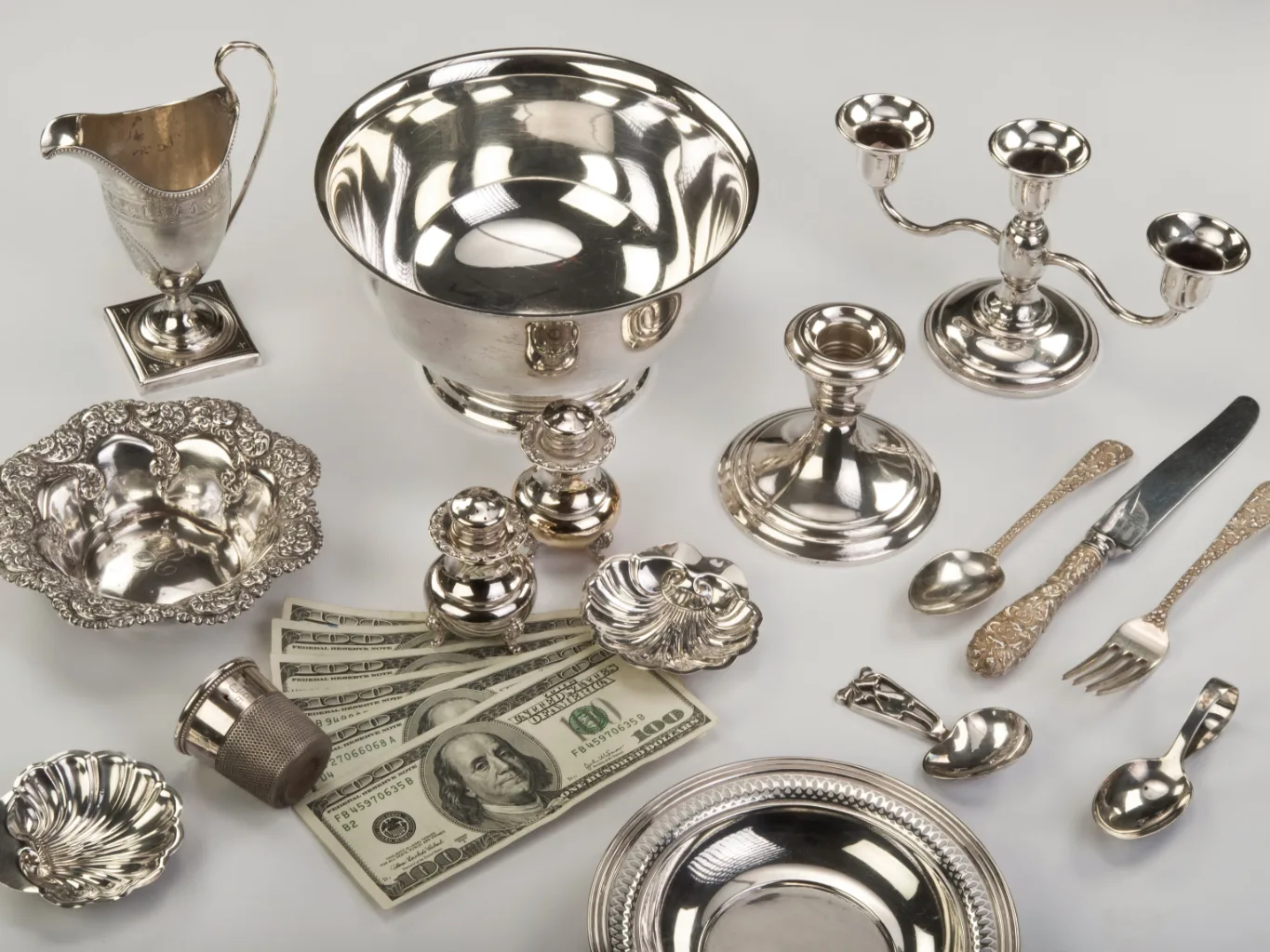 Across the counter in a wide shot, a worker uses a handheld magnifier to inspect a silver pendant during the evaluation stage, with other silver items arranged in a tray nearby